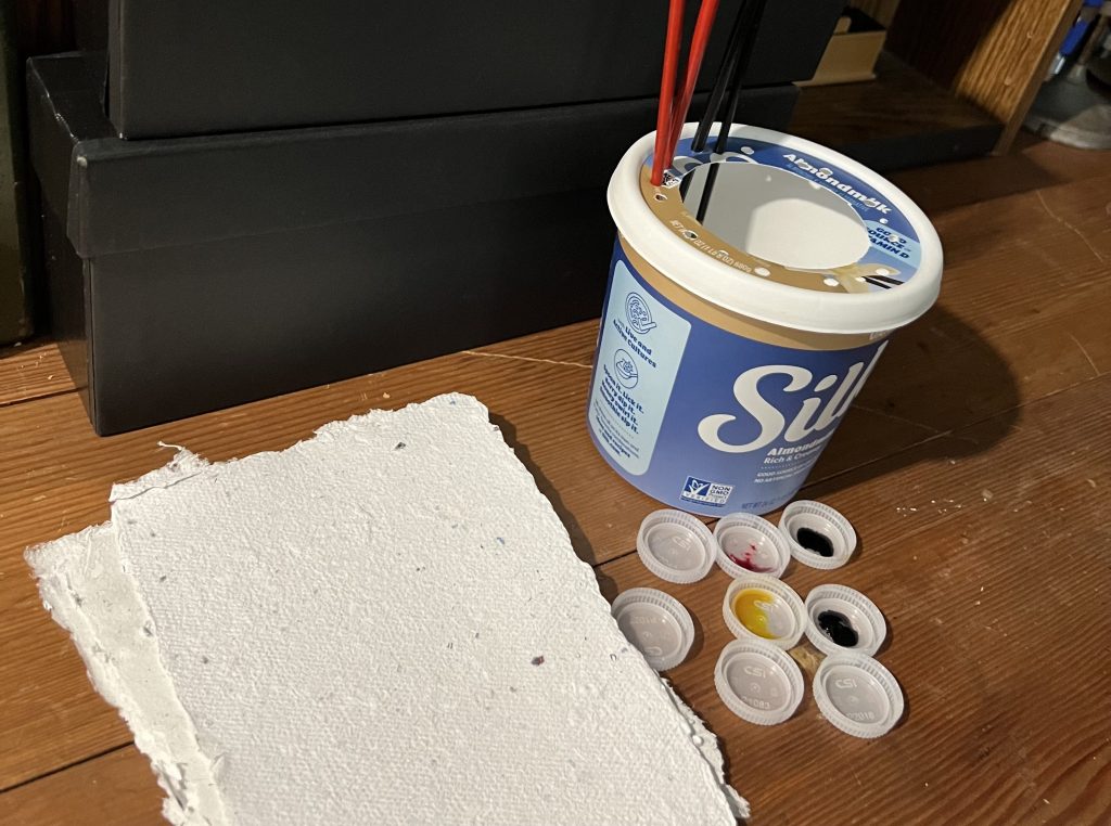 Plastic containers from food being used as paint brush holder water cup, bottle lids as paint wells, and handmade paper from recycled junk mail